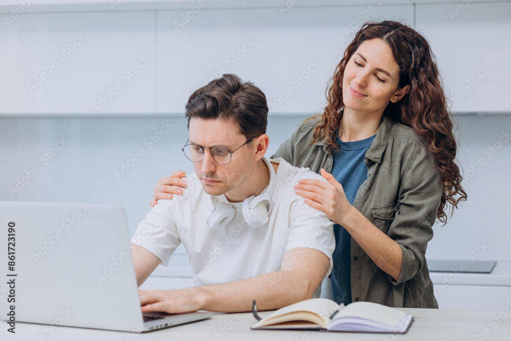 Woman places her hands on man shoulders, providing comfort and support as he focuses on his laptop, indicating a moment of care. Husband works remotely online from home kitchen. Woman places her hands on man shoulders, providing comfort and support as he focuses on his laptop, indicating a moment of care. Husband works remotely online from home kitchen.