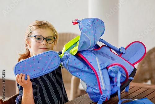 Happy smiling girl preparing for school her backpack. First day of school. Back to school concept. Little child collecting different supplies like pens and books in a bag or satchel.