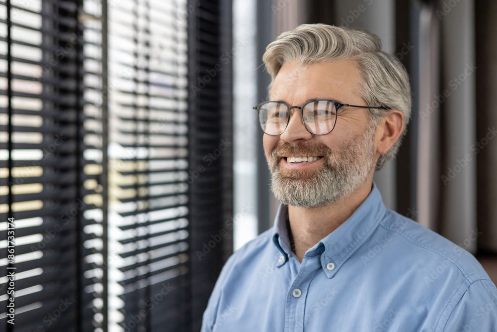 Mature man with glasses smiling while looking out window with blinds in contemporary office environment. Natural light illuminating cheerful expression capturing professional yet relaxed demeanor