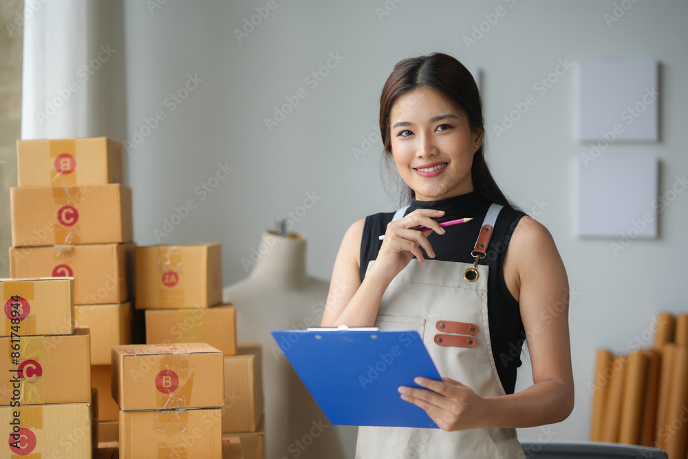 Asian sme business woman holding pencil and clipboard checking order from customer on website in warehouse Asian sme business woman holding pencil and clipboard checking order from customer on website in warehouse