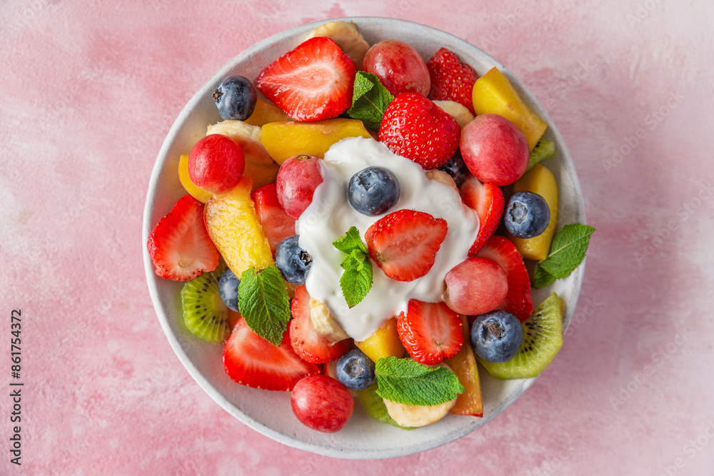 Bowl of fresh fruit salad topped with yogurt and garnished with mint leaves, featuring strawberries, blueberries, kiwi, grapes, banana and peaches on pink background. Top view. Healthy summer food Bowl of fresh fruit salad topped with yogurt and garnished with mint leaves, featuring strawberries, blueberries, kiwi, grapes, banana and peaches on pink background. Top view. Healthy summer food