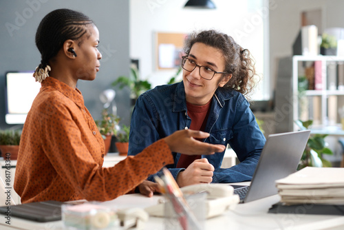 Portrait of smiling young man with long hair listening to female colleague discussing ideas during meeting at workplace in office