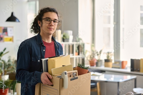 Waist up portrait of young man carrying box with personal items in office and looking at camera with smile copy space