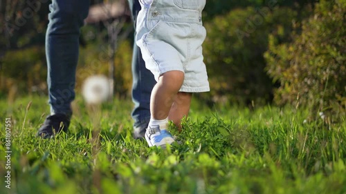 Father helps child take steps on grass. Child learning to walk outdoors. Happy bonding time with father. First steps in nature. Step, child, grass father moment. Happy family in the park. Happy family photo