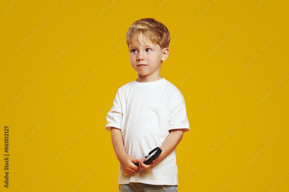 Thoughtful little boy in white tshirt looking away while holding in hands a magnifier, isolated against yellow background. Thoughtful little boy in white tshirt looking away while holding in hands a magnifier, isolated against yellow background.