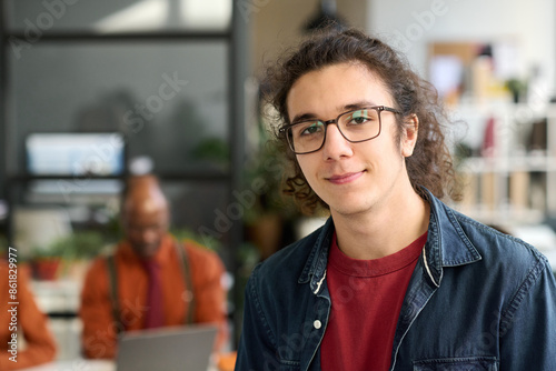 Candid close up portrait of long haired young man wearing glasses looking at camera in office and smiling internship concept copy space © Mediaphotos