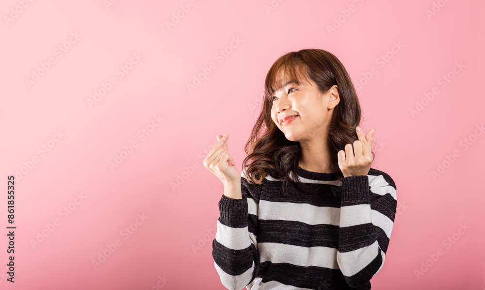 A confident woman stands with a smile, creating a mini heart sign with her finger on a pink background. Asian portrait of a beautiful young female sending love for Valentine's Day in a studio shot. A confident woman stands with a smile, creating a mini heart sign with her finger on a pink background. Asian portrait of a beautiful young female sending love for Valentine's Day in a studio shot.
