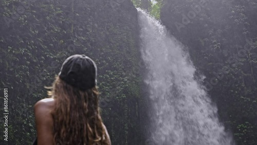 A woman in a black outfit and cap gazes at a majestic waterfall surrounded by lush greenery. The serene scene captures the beauty of nature and the power of the waterfall.