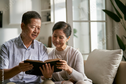 senior asian couple sitting on couch at home reading the bible together