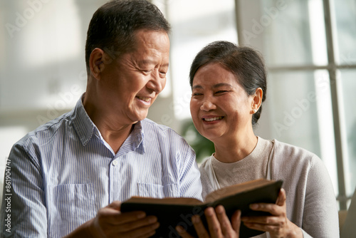senior asian couple sitting on couch at home reading bible together with smile and happiness