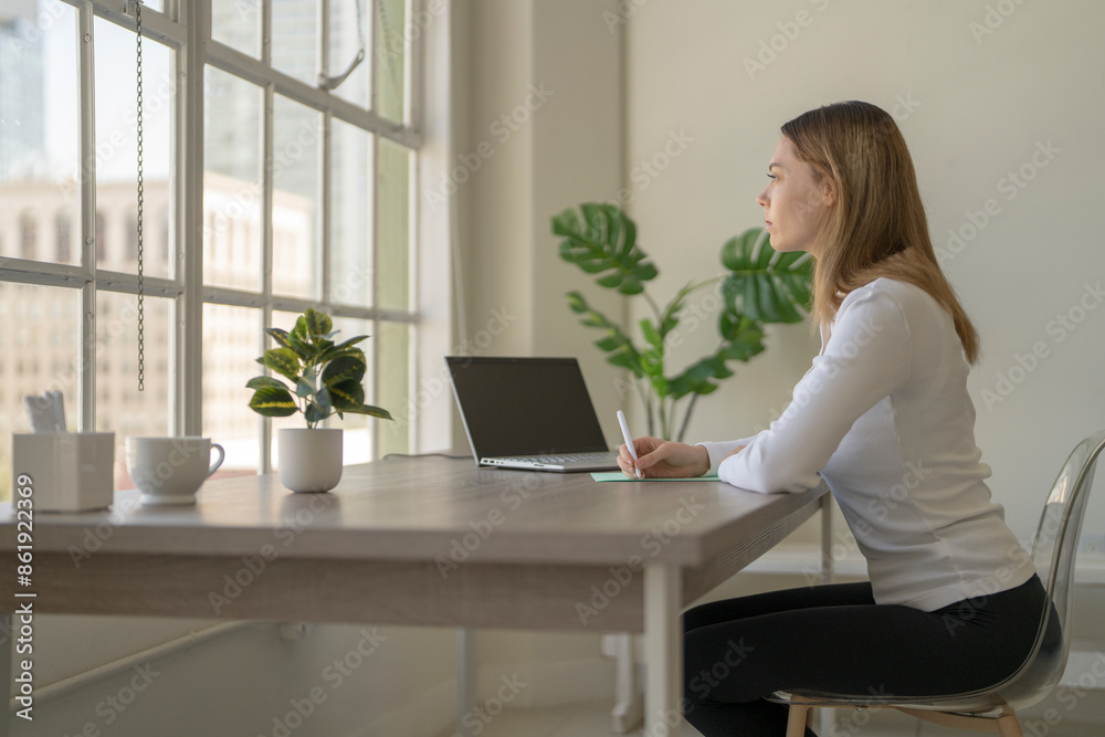 Gen Z female with long brunette office worker at desk writing notes, looking out of a large bright window Gen Z female with long brunette office worker at desk writing notes, looking out of a large bright window