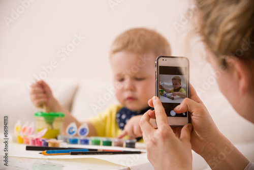 Wallpaper Mural Young mother taking photo of cute little toddler boy in yellow shirt sitting at table and drawing with colorful paints. Early learning. Creative. Toddler drawing. Mom making picture on mobile phone Torontodigital.ca