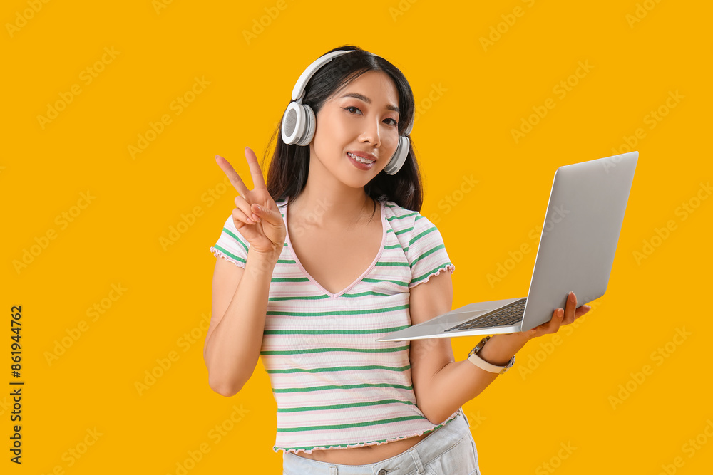 Young female Asian programmer in headphones with laptop showing thumb-up gesture on yellow background Young female Asian programmer in headphones with laptop showing thumb-up gesture on yellow background