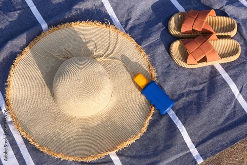 Towel with stylish straw hat, bottle of sunscreen cream and flip-flops outdoors