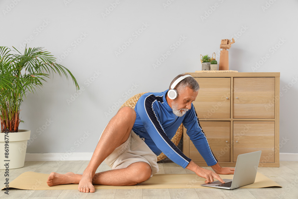 Fototapeta premium Sporty mature man in headphones using laptop on mat at home