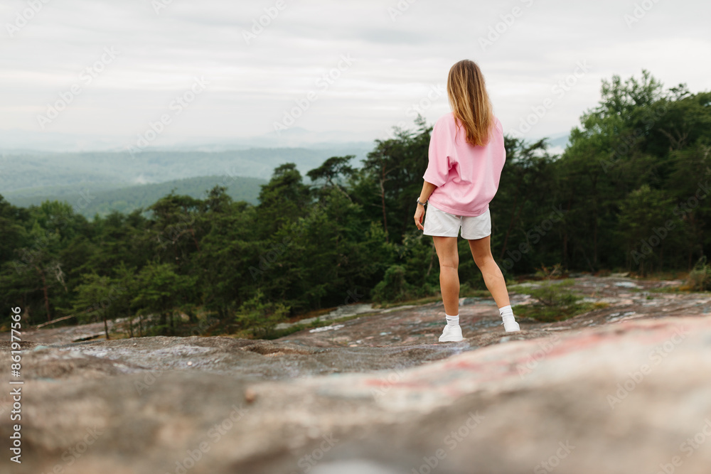 A young girl in white shorts, a pink T-shirt and white sneakers stands on a rock and looks at the mountains on a summer day. Dawn in the mountains. Woman watching the sunrise in the mountains. Hiker A young girl in white shorts, a pink T-shirt and white sneakers stands on a rock and looks at the mountains on a summer day. Dawn in the mountains. Woman watching the sunrise in the mountains. Hiker