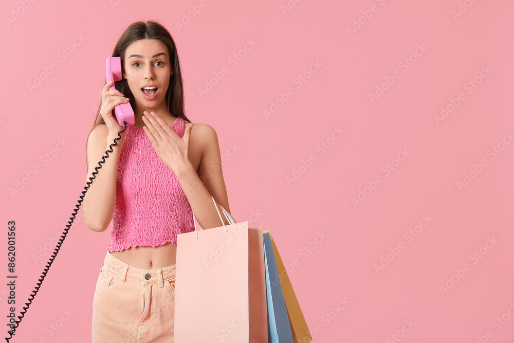 Shocked young woman talking by telephone with shopping bags on pink background