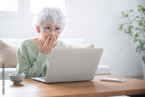 パソコンを見ながら詐欺サイトや何かフィッシングメールで口に手を当てる驚くシニア女性 ネガティブなイメージ
