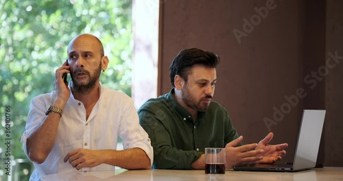Two handsome middle-aged businessmen are having a meeting on their laptops outside the office environment. One of the men ignores the discussion and takes a phone call, which makes the other man angry