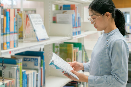 A woman wearing glasses is reading a book in a library next to a bookshelf.