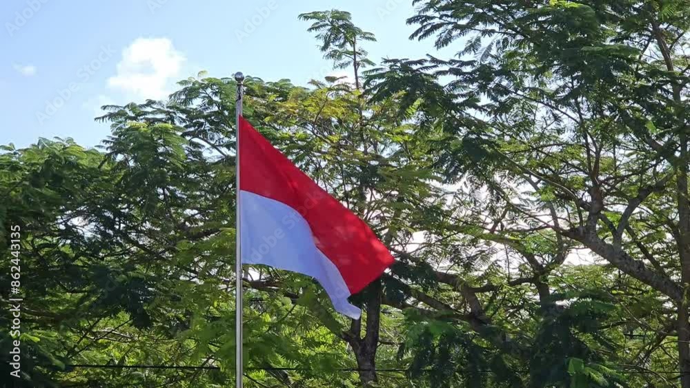 The Indonesian flag flutters in the wind on a pole with a blue sky and greenery background