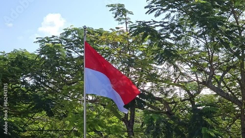 The Indonesian flag flutters in the wind on a pole with a blue sky and greenery background photo