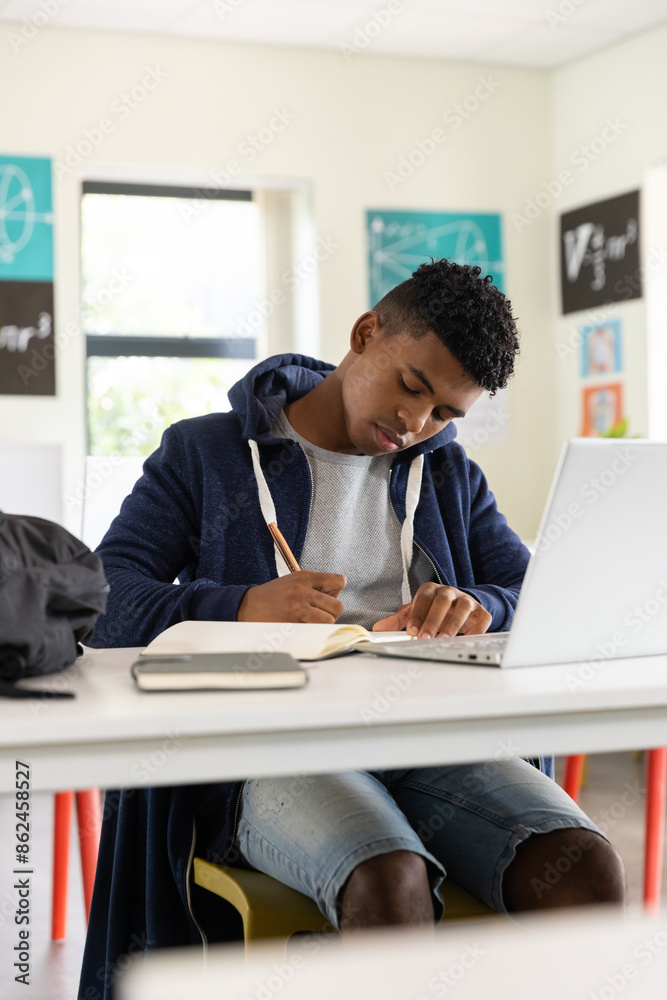 Writing in notebook and using laptop, African American male student in school classroom Writing in notebook and using laptop, African American male student in school classroom