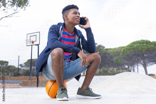 Sitting on basketball, African American teenage boy talking on smartphone outdoors