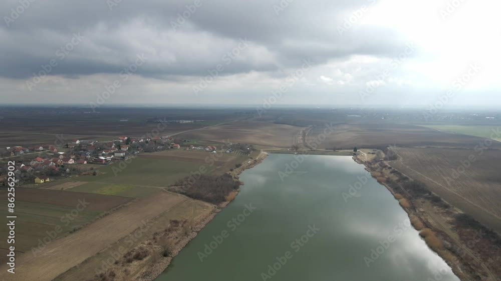 Drone captures a lake surrounded with fields and village under cloudy moody sky Drone captures a lake surrounded with fields and village under cloudy moody sky