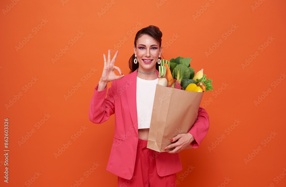 friendly smiling woman holding paper shopping bag full of groceries isolated on orange studio shot background. friendly smiling woman holding paper shopping bag full of groceries isolated on orange studio shot background.
