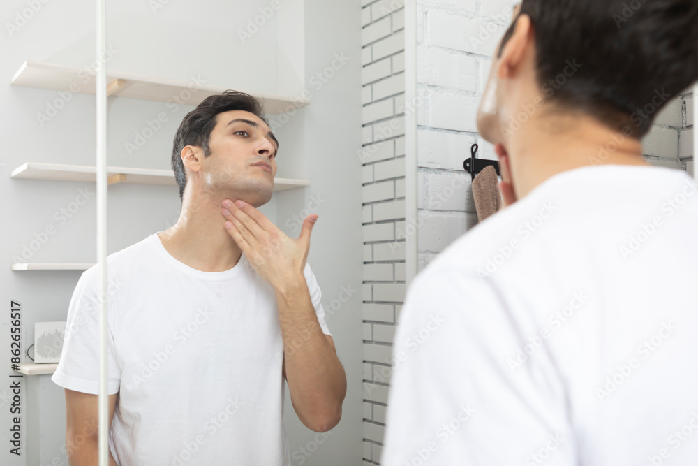 Handsome man looking at mirror and applying moisturizing cream on cheeks in bathroom. Groomed young guy doing skincare morning routine after taking a shower Handsome man looking at mirror and applying moisturizing cream on cheeks in bathroom. Groomed young guy doing skincare morning routine after taking a shower