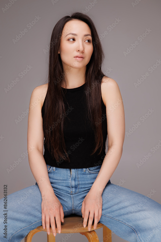 Asian woman in black top and blue jeans sitting on chair isolated on grey Asian woman in black top and blue jeans sitting on chair isolated on grey
