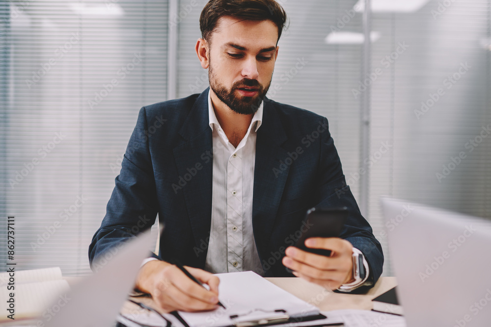 Pensive proud ceo rewriting information data from smartphone device working in office interior.Bearded businessman reading incoming notification on telephone while making accounting reports Pensive proud ceo rewriting information data from smartphone device working in office interior.Bearded businessman reading incoming notification on telephone while making accounting reports
