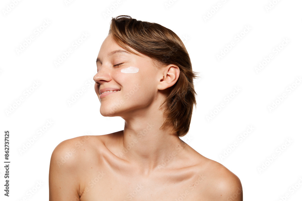 Pretty woman with short brown hair smiles softly with her eyes closed, showing dab of lotion on her cheek against white studio background. Concept of youth, beauty, cosmetic product, dermatology. Ad Pretty woman with short brown hair smiles softly with her eyes closed, showing dab of lotion on her cheek against white studio background. Concept of youth, beauty, cosmetic product, dermatology. Ad