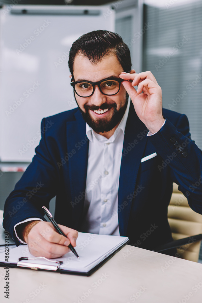 Successful bearded businessman in stylish eyeglasses for vision correction checking accounting reports and noting data while smiling at camera.Positive proud ceo in formal wear writing information Successful bearded businessman in stylish eyeglasses for vision correction checking accounting reports and noting data while smiling at camera.Positive proud ceo in formal wear writing information