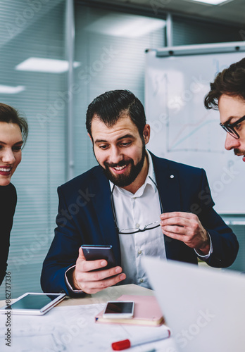 Young good looking skilled smiling colleagues watching together funny video in website on smartphone connected to internet.Group of intelligent emotional architects having fun during work break photo