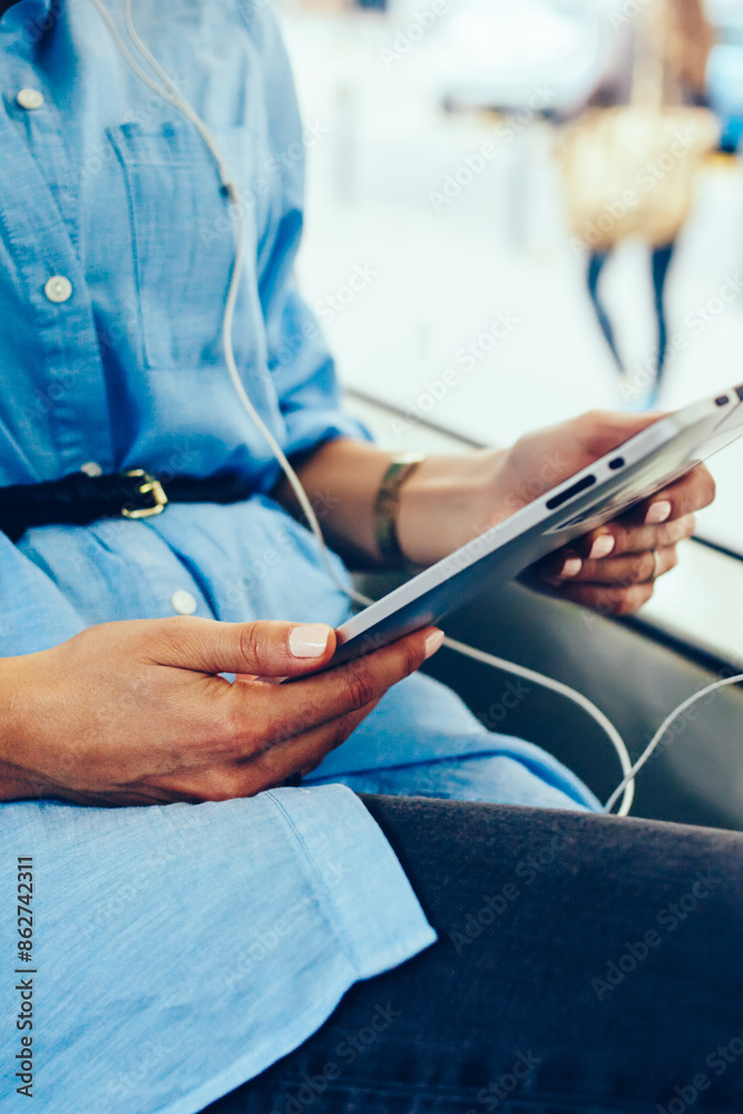 Cropped view of woman's hands holding modern tablet with earphones sitting in tram.Hipster girl listening audio music downloaded in player on touch pad using public wireless internet connection Cropped view of woman's hands holding modern tablet with earphones sitting in tram.Hipster girl listening audio music downloaded in player on touch pad using public wireless internet connection