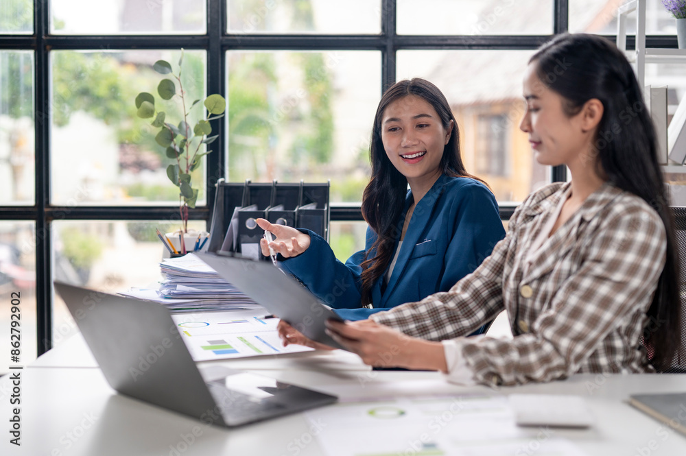 Two women are sitting at a desk with a laptop and papers