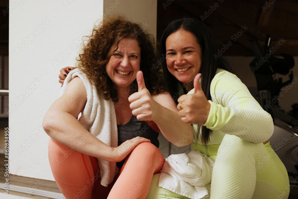 2 Frauen im Fitnessstudio machen Pause, lachend und Daumen hoch 2 Frauen im Fitnessstudio machen Pause, lachend und Daumen hoch