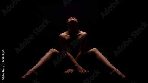 A woman sits in a dark room, her head tilted back and her legs spread out. She is bare-chested, and the light from above illuminates her face and body.