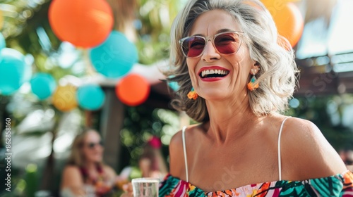 Smiling senior woman with gray hair and sunglasses enjoying a summer party outdoors, surrounded by colorful decorations and people in the background