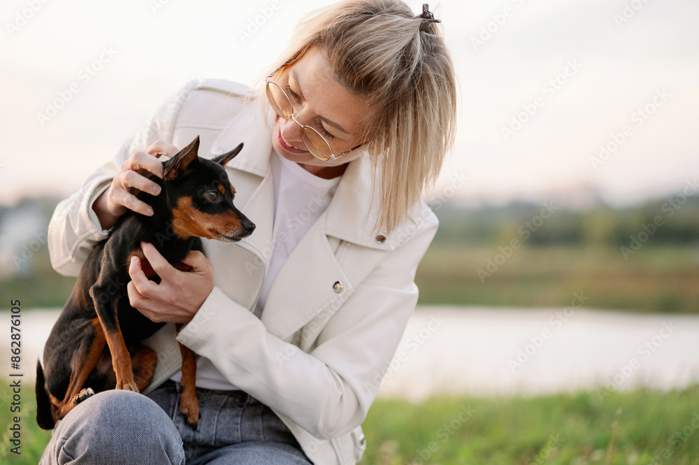 Woman smiles while holding small smooth-haired Russian toy terrier dog in her arms while walking. Friendship between animals and humans, caring for pet Woman smiles while holding small smooth-haired Russian toy terrier dog in her arms while walking. Friendship between animals and humans, caring for pet