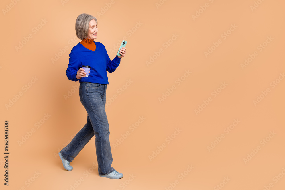 Full length photo of lovely pensioner lady walk hold device coffee dressed stylish blue garment isolated on beige color background Full length photo of lovely pensioner lady walk hold device coffee dressed stylish blue garment isolated on beige color background