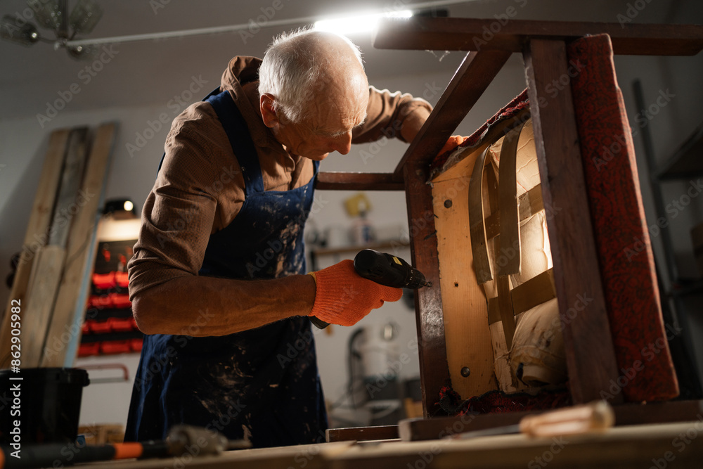elderly man in orange gloves and a blue apron repairs a chair with an electric drill in a workshop. He works on a wooden frame, dismantling the old structure. elderly man in orange gloves and a blue apron repairs a chair with an electric drill in a workshop. He works on a wooden frame, dismantling the old structure.