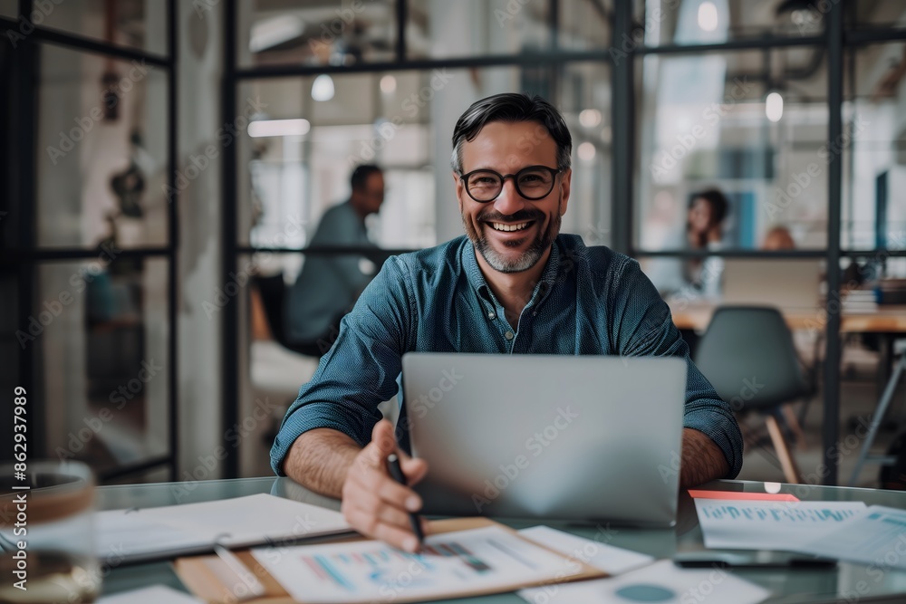A smiling man in glasses sitting at desk with laptop holding pen looking into camera Professional stress free Workspace Concept A smiling man in glasses sitting at desk with laptop holding pen looking into camera Professional stress free Workspace Concept