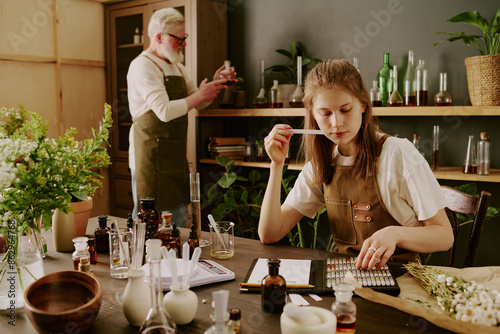Female perfumer sitting at table and smelling tester while mature male perfumer reading notes