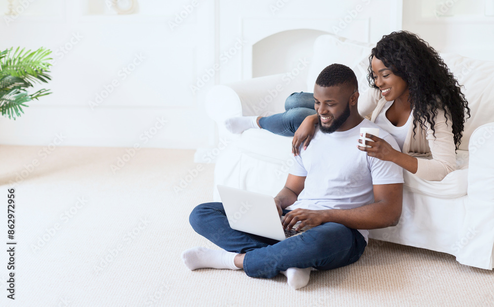 A couple relaxes on the floor with a laptop. The man is sitting on the floor, while the woman is sitting on the couch next to him. They both look happy and relaxed. A couple relaxes on the floor with a laptop. The man is sitting on the floor, while the woman is sitting on the couch next to him. They both look happy and relaxed.