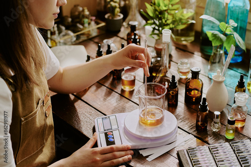 Young female perfumer blending essential oils while she weighing it with plastic kitchen scale