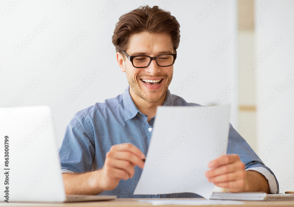 A man wearing glasses smiles and laughs while reading a document at his desk.