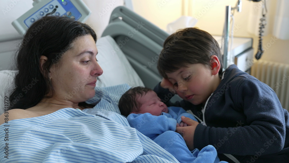 Newborn baby's first day of life welcomed by brother at hospital clinic, infant laid in bed besides mother and sibling. Authentic real life heartwarming family scene, maternal carE Newborn baby's first day of life welcomed by brother at hospital clinic, infant laid in bed besides mother and sibling. Authentic real life heartwarming family scene, maternal carE